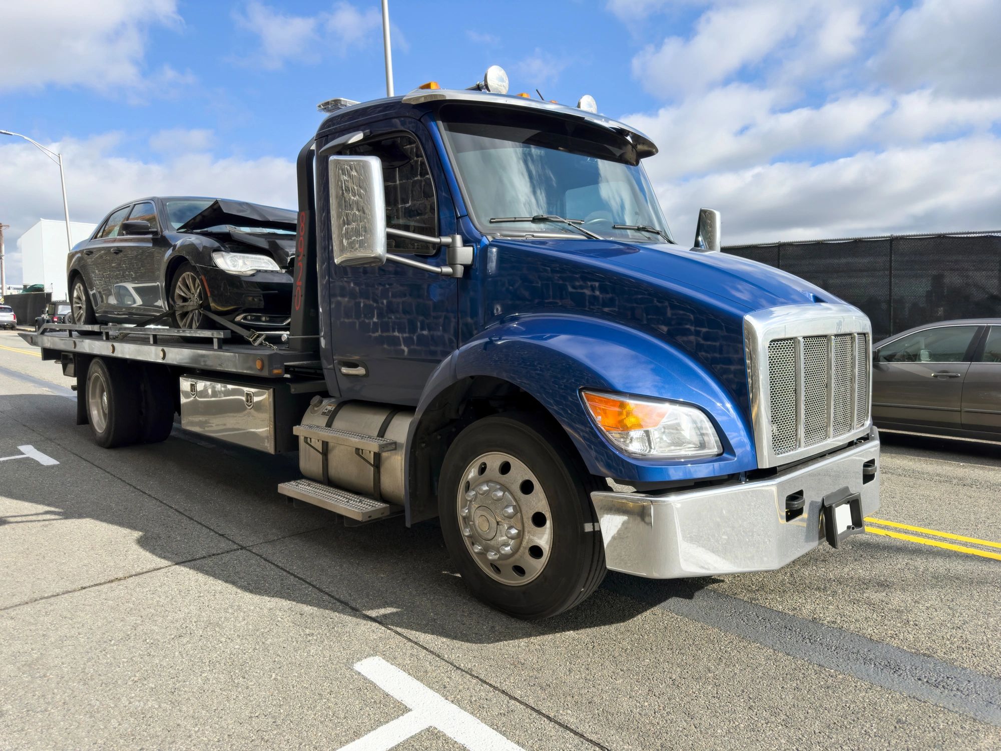 Blue flatbed tow truck carrying a black sedan with front-end damage