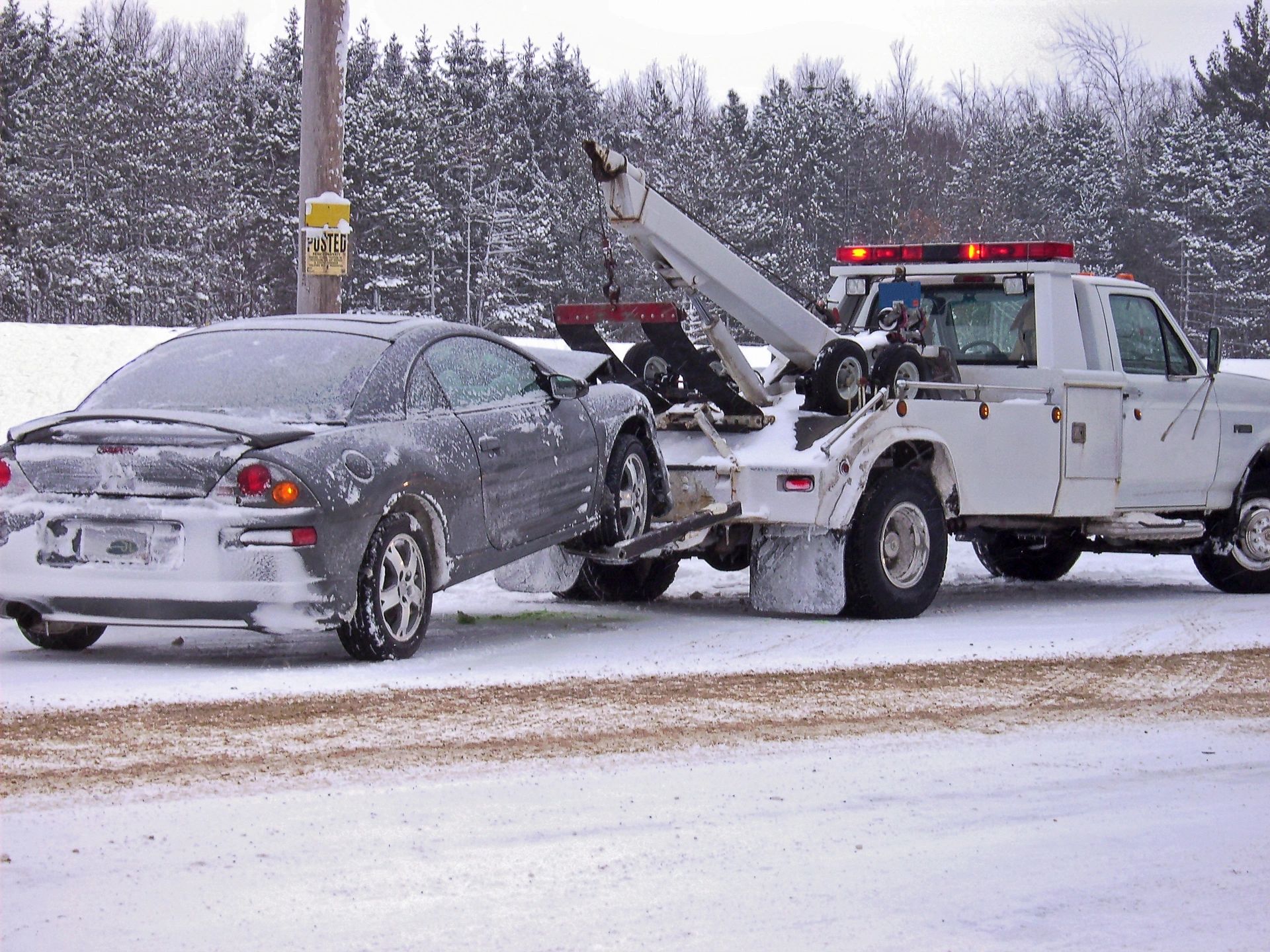 White tow truck with flashing red lights towing a snow-covered car on a snowy road
